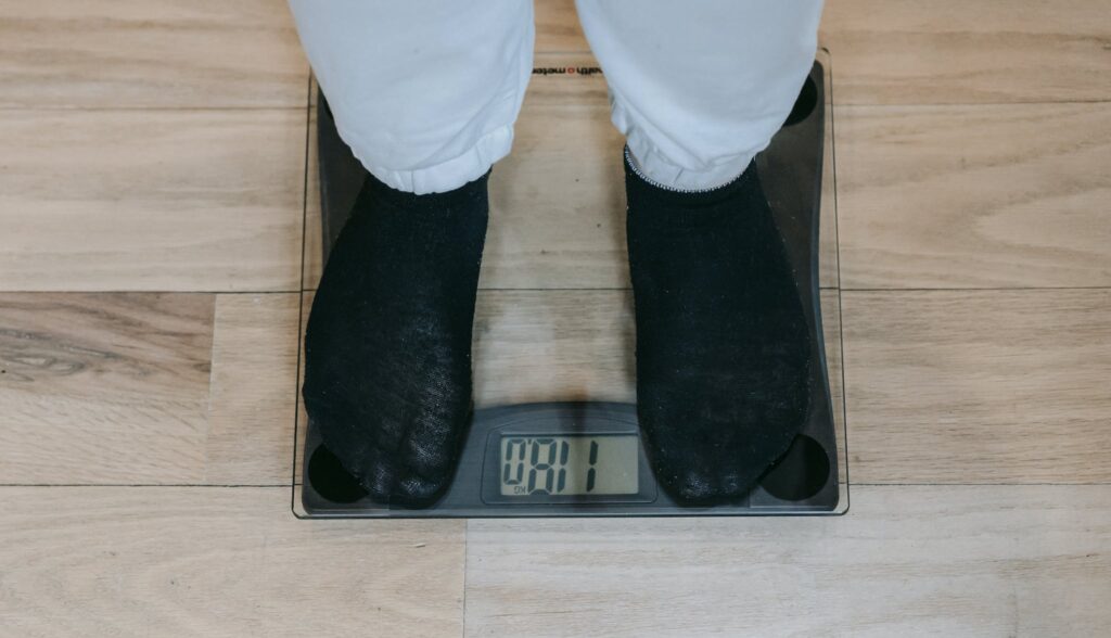 From above of crop unrecognizable plus size person in casual clothes standing on weighing scale on wooden floor